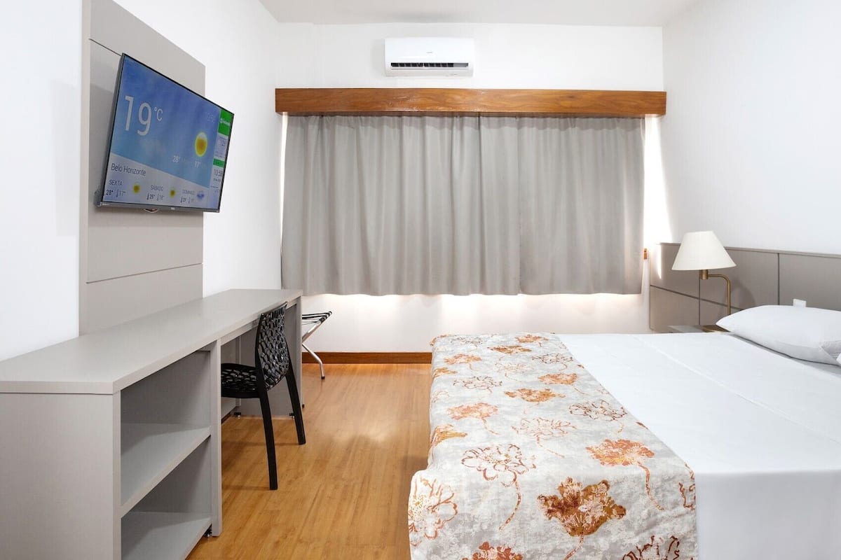 Hotellihuone, jossa on sänky, työpöytä, tuoli, televisio, jossa näkyy huoneen lämpötila, ja ilmastointilaite.