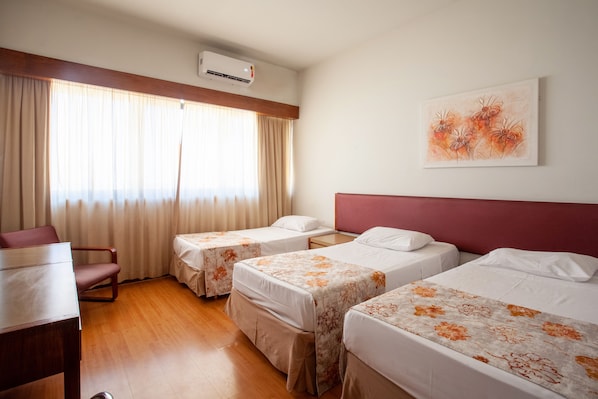 A hotel room with two beds, a desk, and a chair. There is an air conditioning unit on the wall and a painting above the beds.
