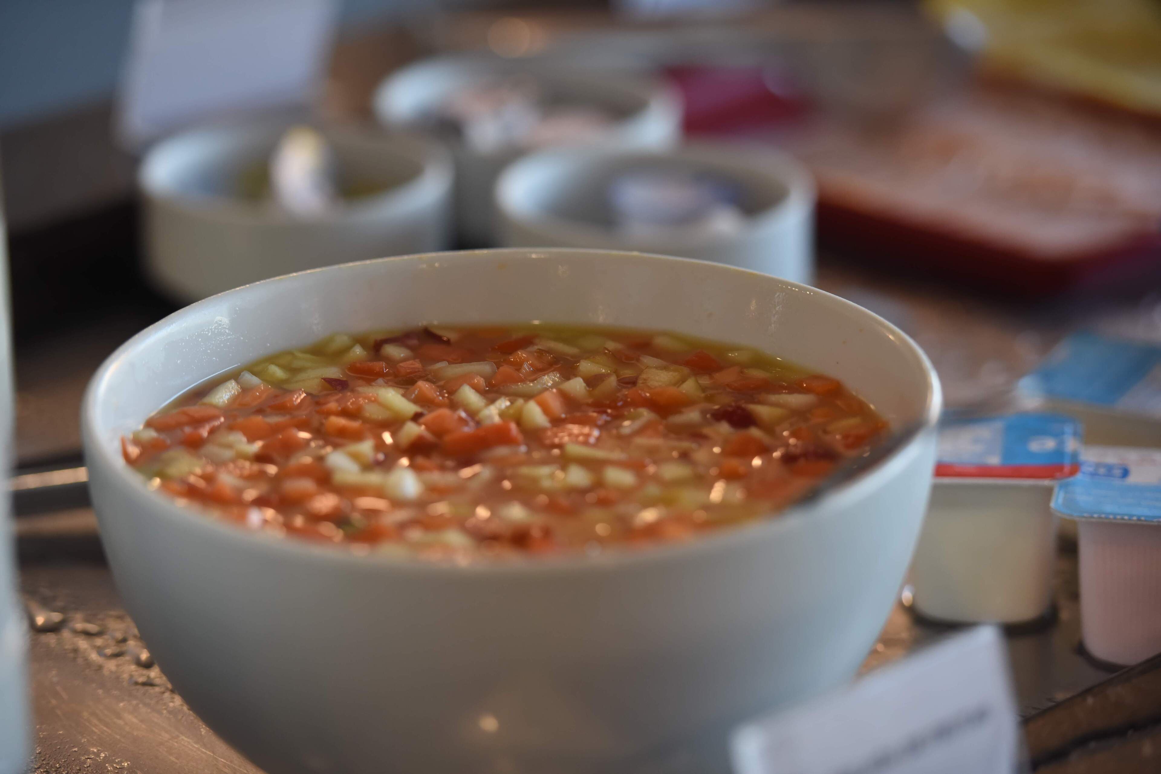 Un tazón de sopa con ingredientes visibles como tomates y cebollas, sobre una mesa con un fondo desenfocado.