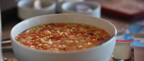 Un tazón de sopa con ingredientes visibles como tomates y cebollas, sobre una mesa con un fondo desenfocado.