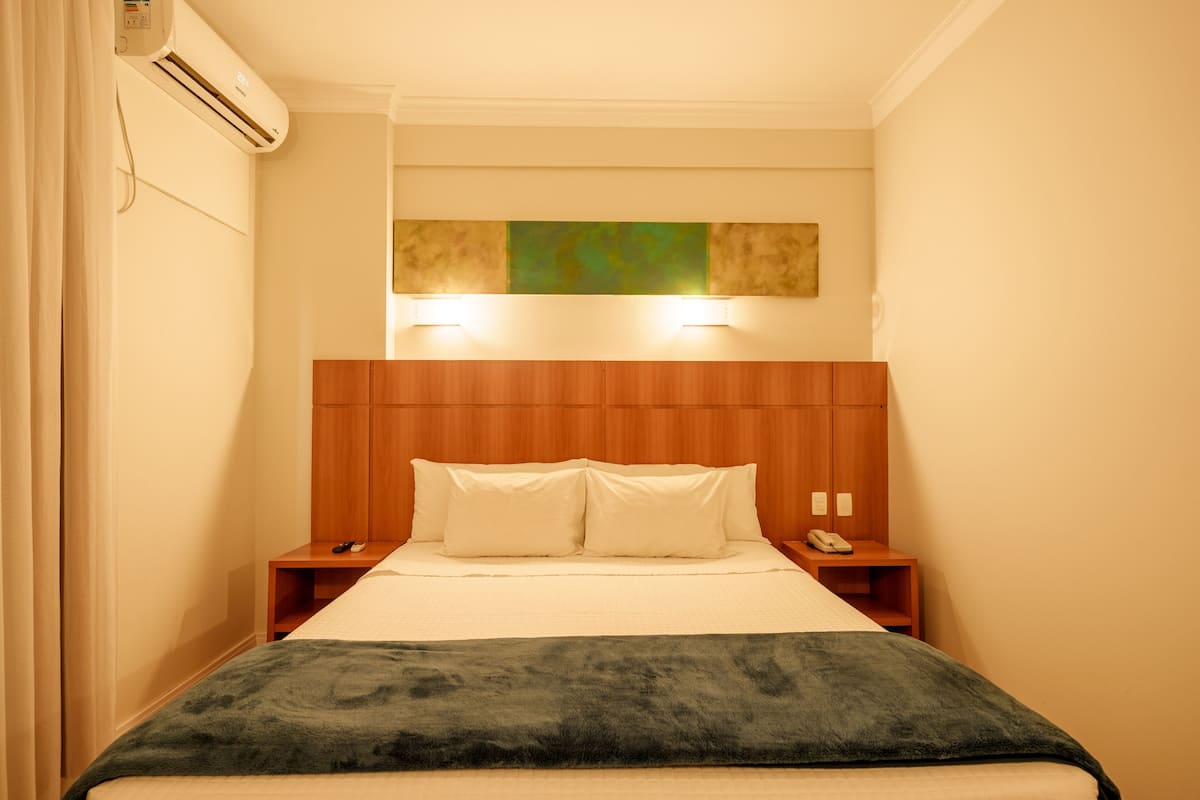 A hotel room with a wooden headboard, a bed with white linens, two bedside tables with phones, and a wall-mounted air conditioner.