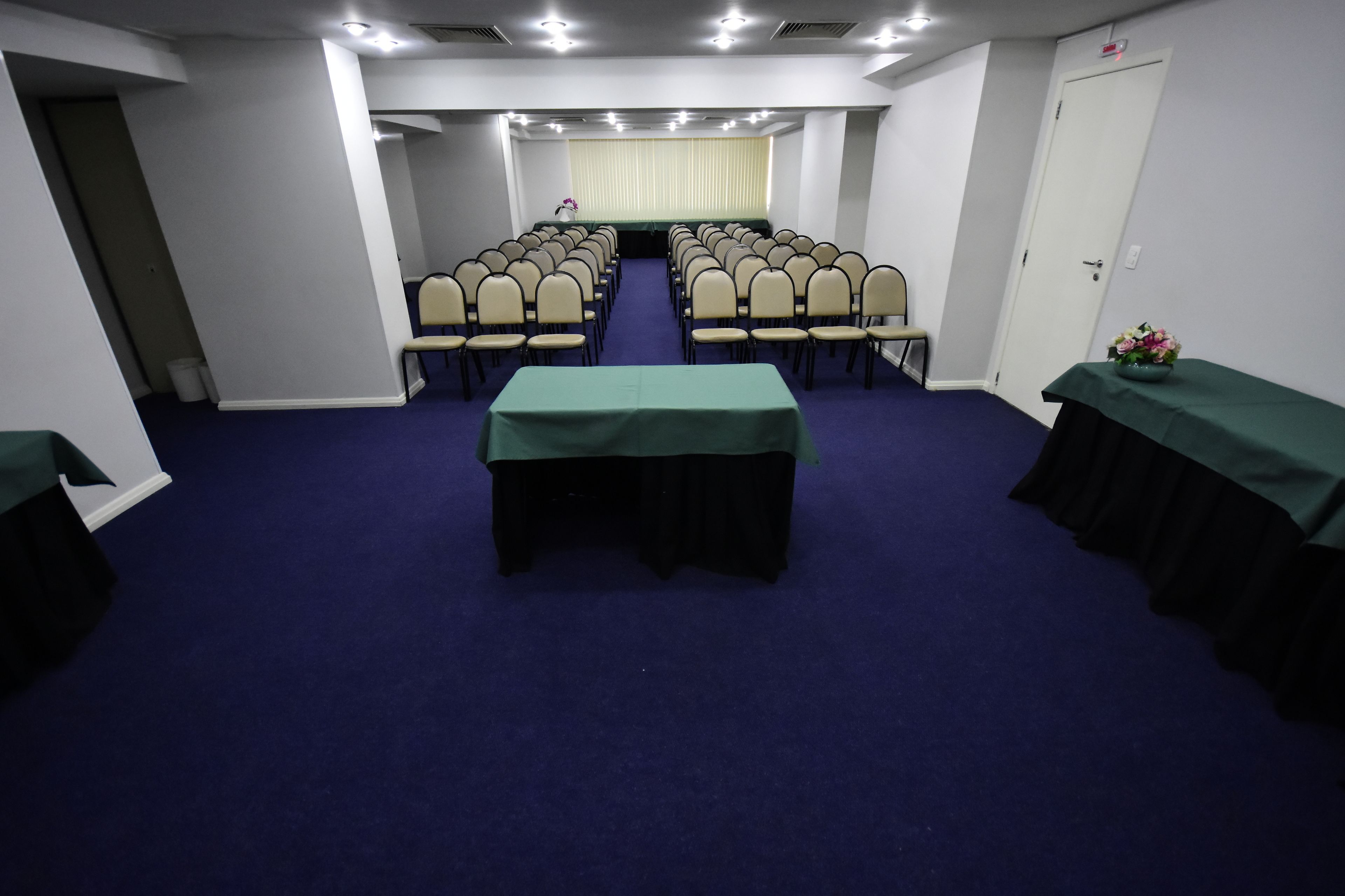 Un salón de conferencias con filas de sillas, una mesa con mantel verde y una puerta a la derecha.