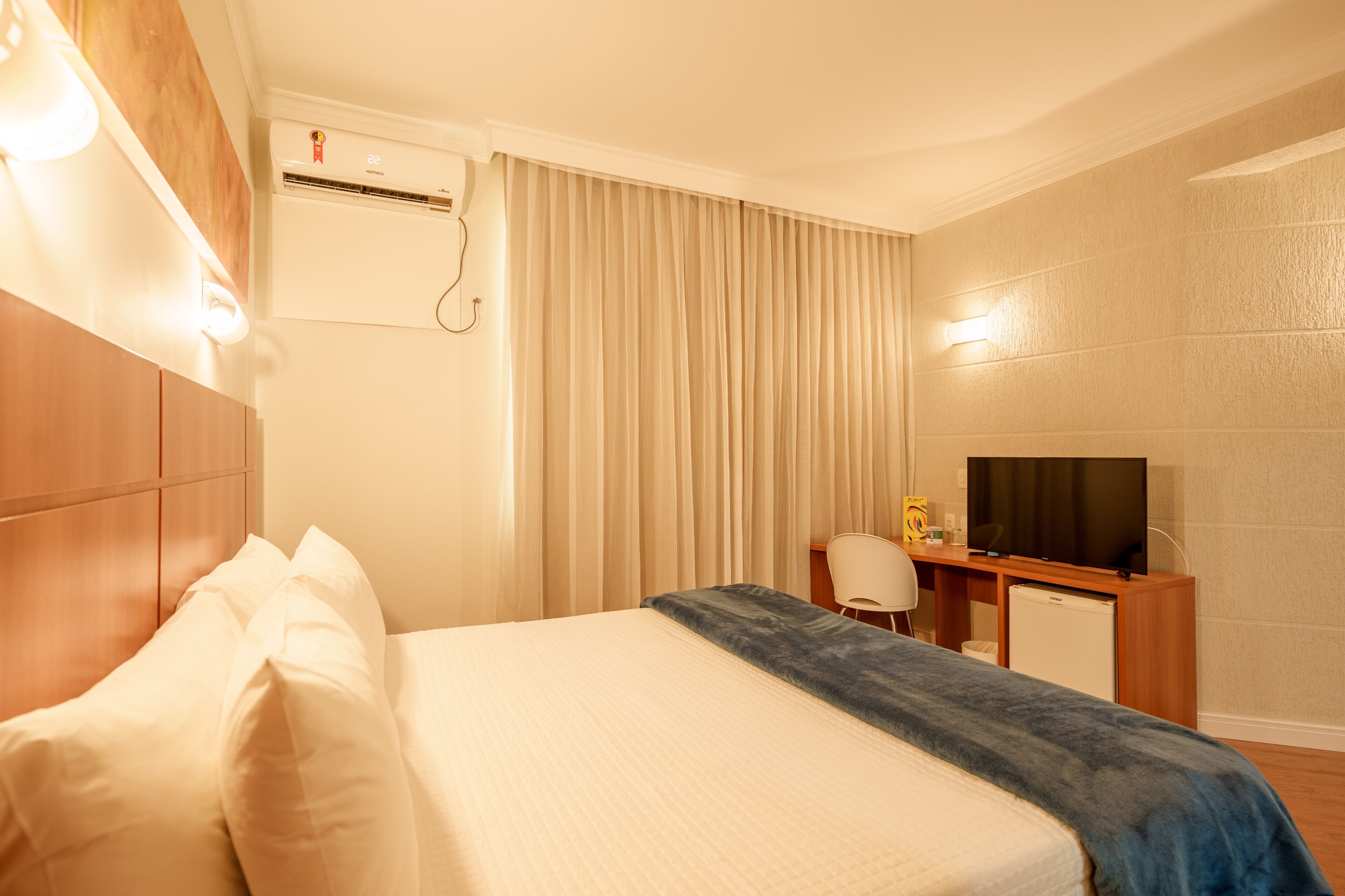 A hotel room with a bed, a desk, a television, and an air conditioning unit.