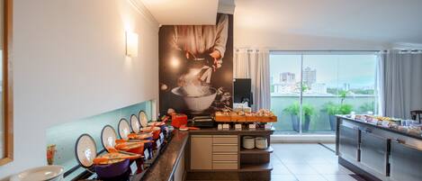A modern hotel kitchen with a buffet setup, featuring a variety of dishes and a large window with a city view.