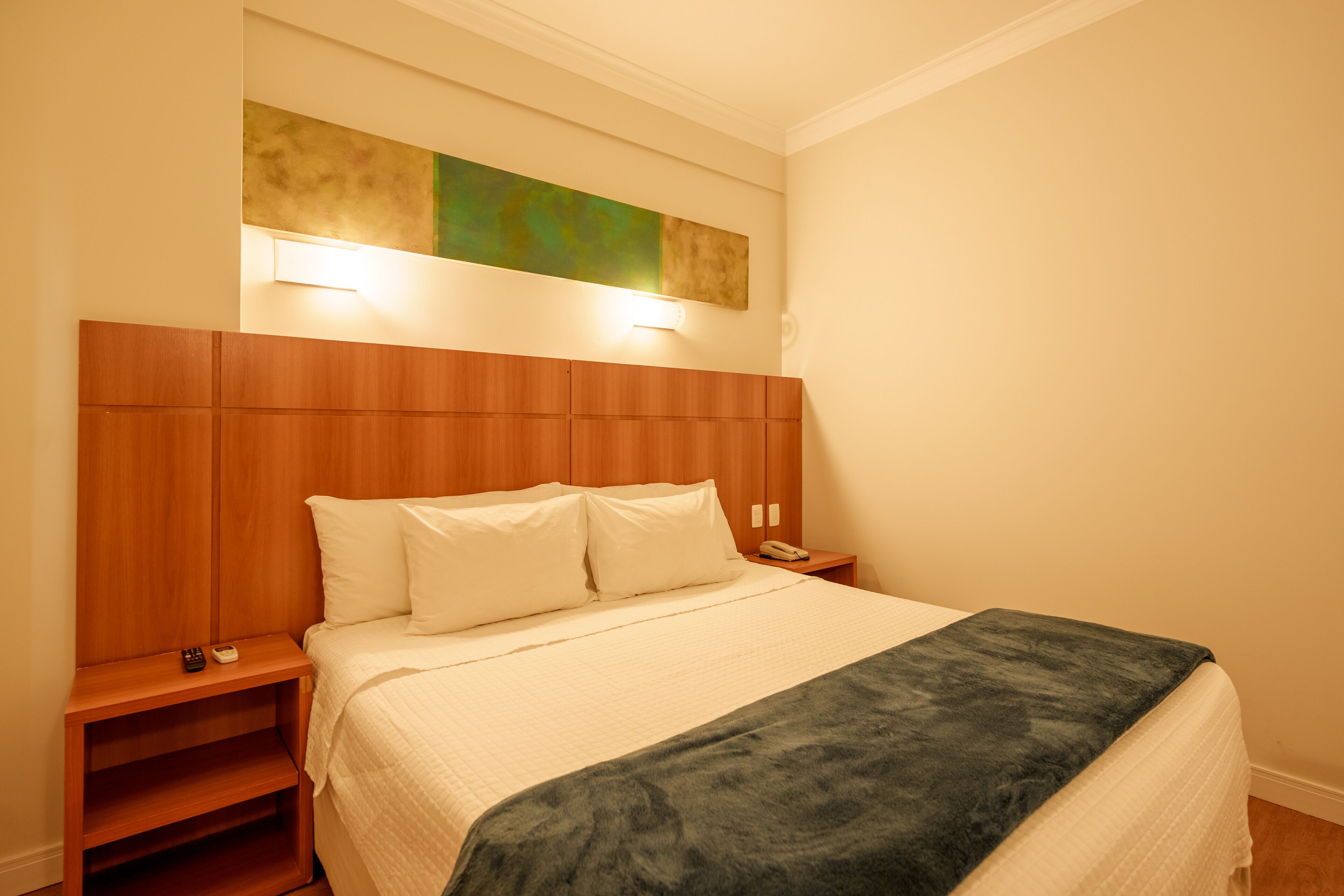 Uma cama bem arrumada com cabeceira de madeira, mesa de cabeceira com abajur e uma obra de arte na parede acima da cama.