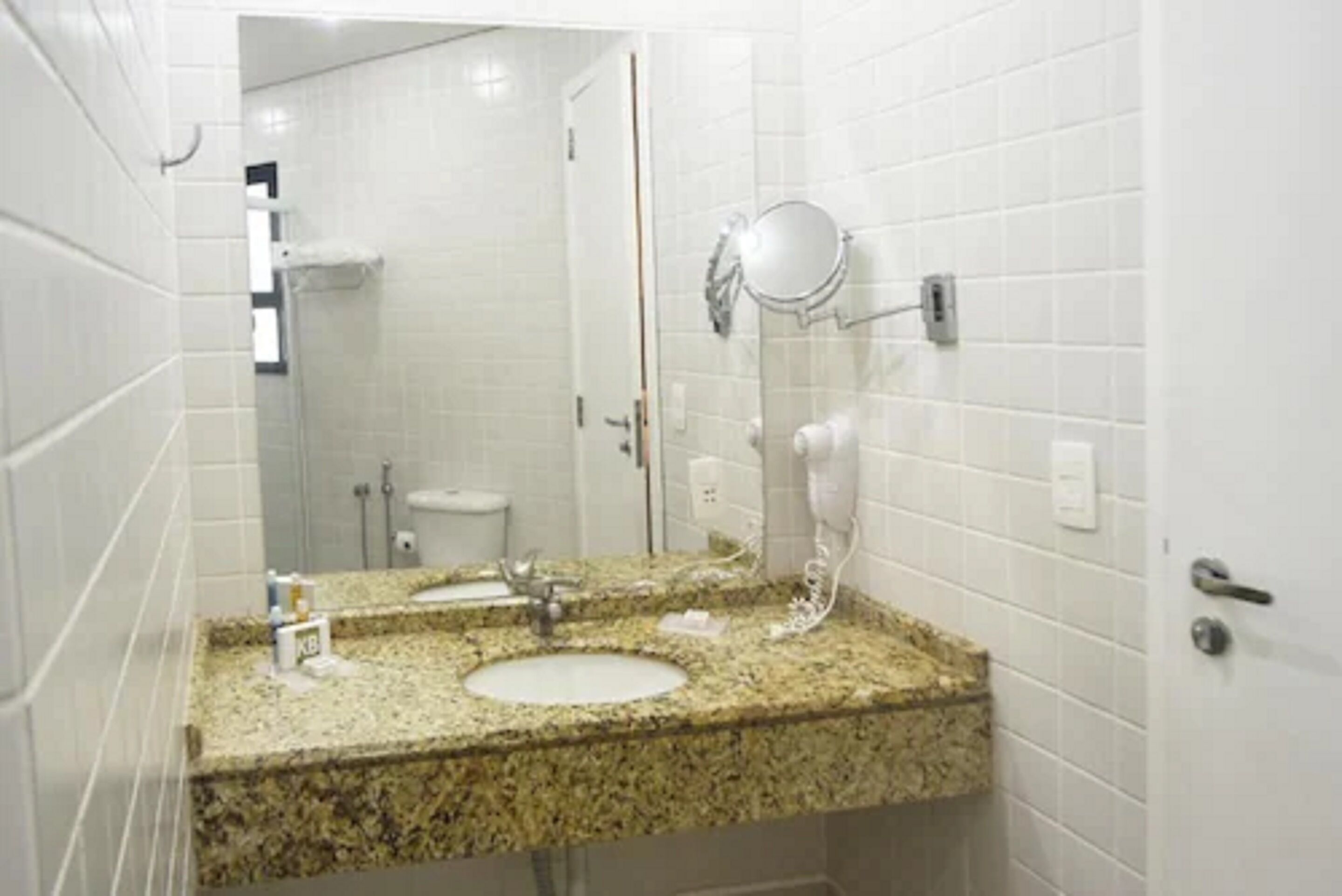 Une salle de bain avec des murs recouverts de carreaux blancs, un comptoir en granit, un lavabo, un miroir et un sèche-cheveux.