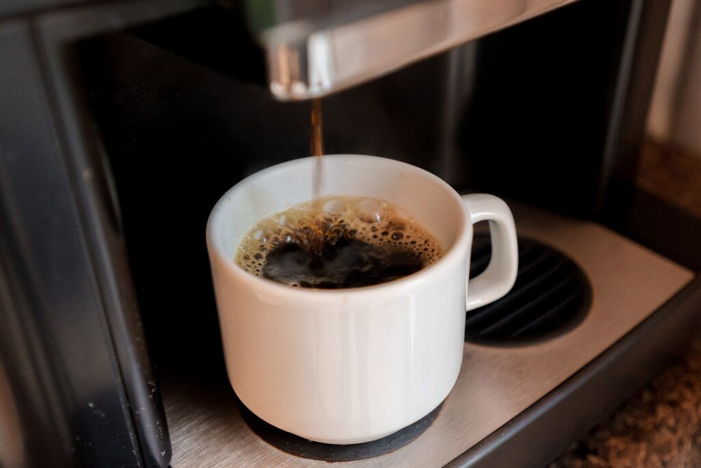A white cup on a metallic tray with coffee being dispensed into it.