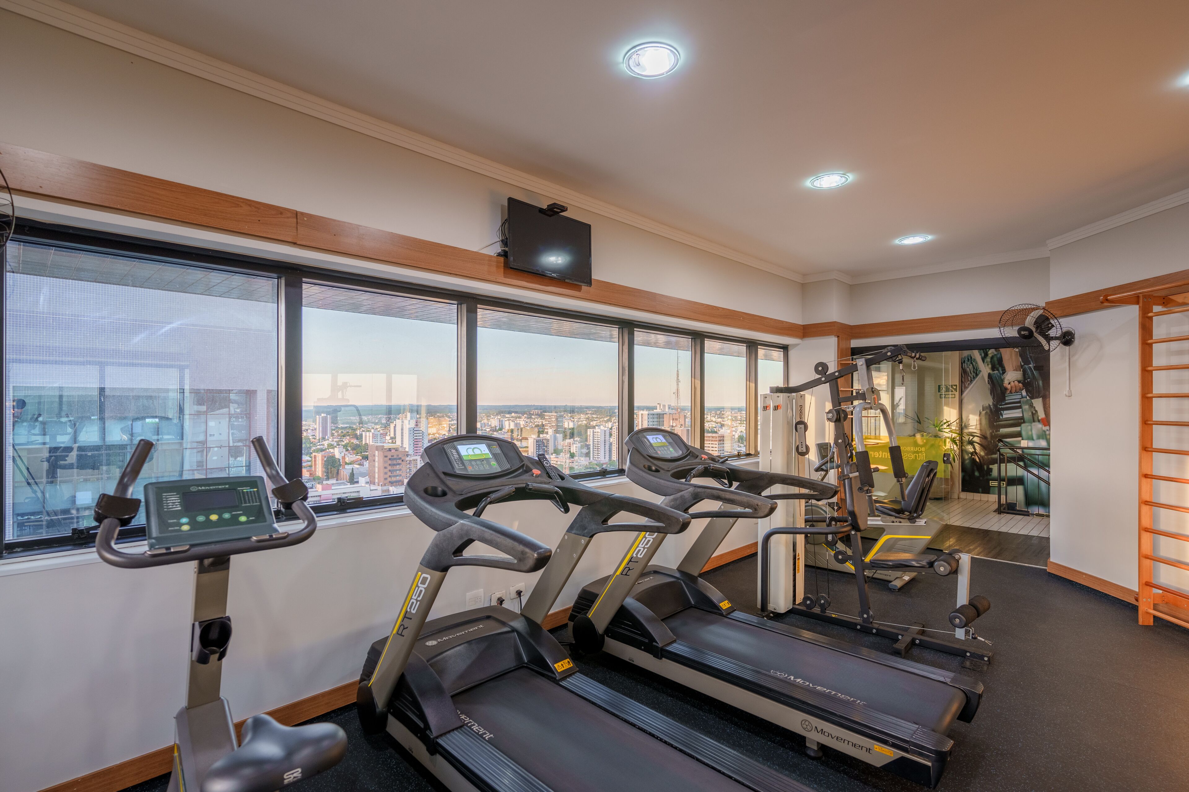 A gym with treadmills, exercise bikes, and weight machines, large windows with a city view, and a wooden ceiling.