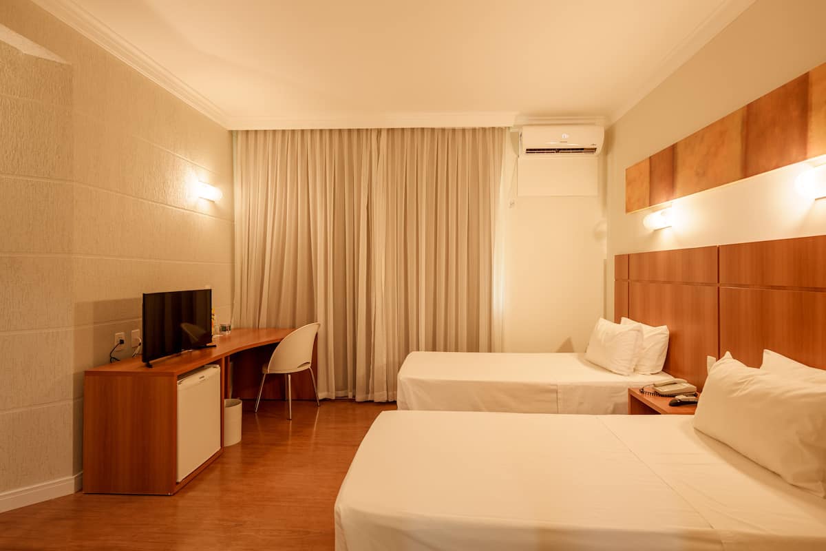 A hotel room with two beds, a desk, a chair, a television, and an air conditioning unit.
