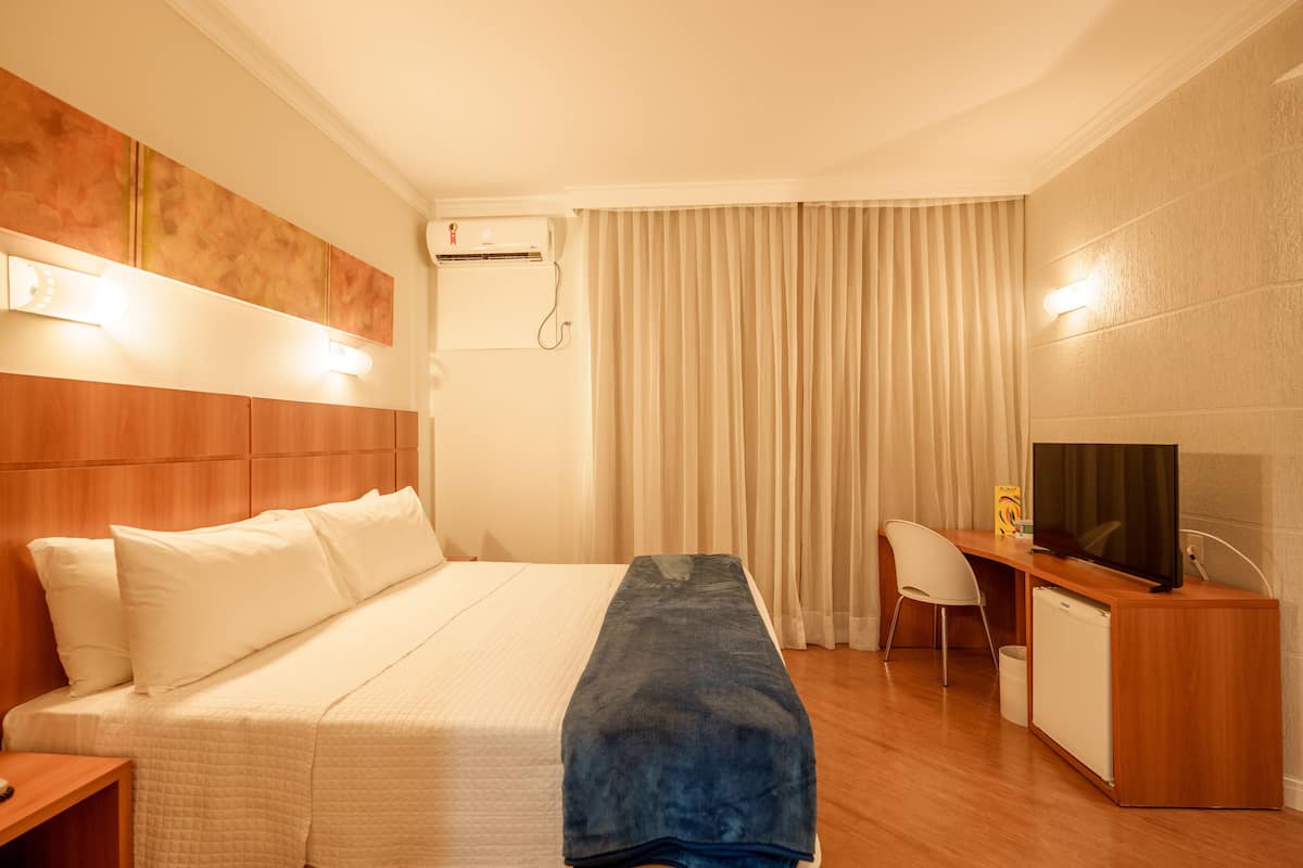A hotel room with a bed, a desk with a television, a chair, and a small refrigerator.