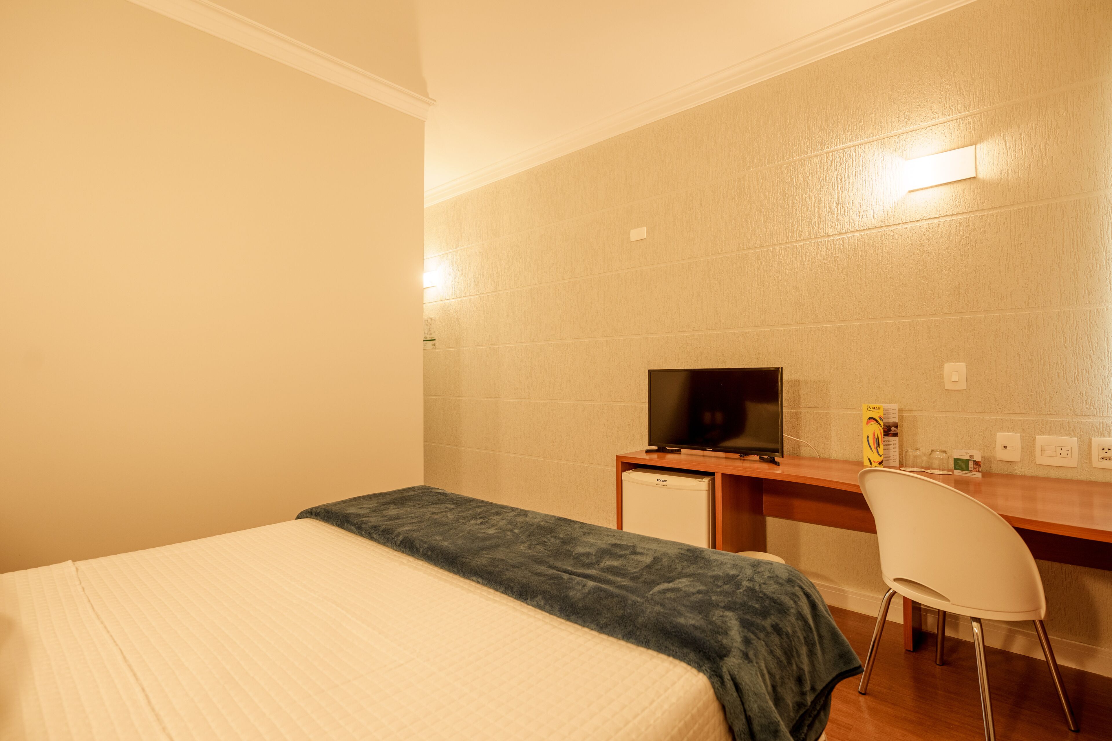 Una habitación de hotel con una cama, un escritorio con televisor, una silla y un refrigerador pequeño.