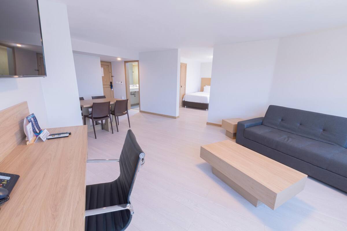 A hotel room with a bed, a sofa, a desk with a computer, a chair, and a dining area with a table and chairs.