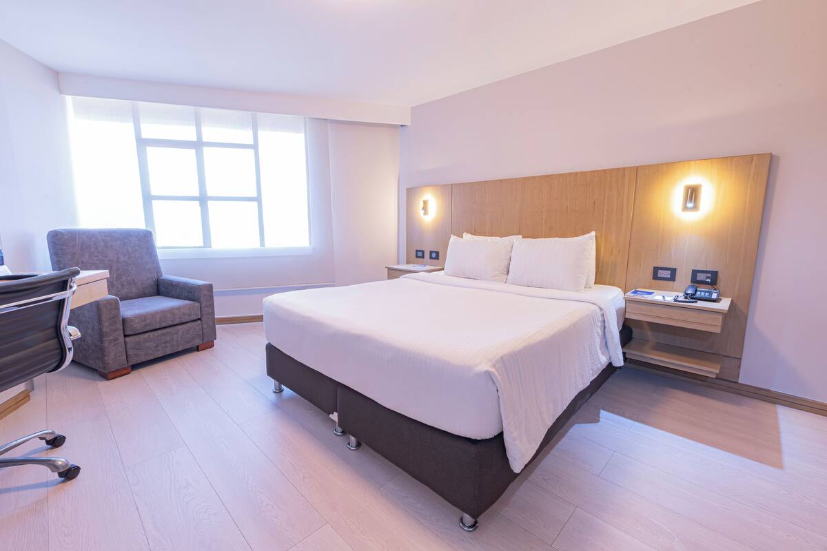 A modern hotel room with a large bed, a desk, a chair, and a nightstand with a lamp.