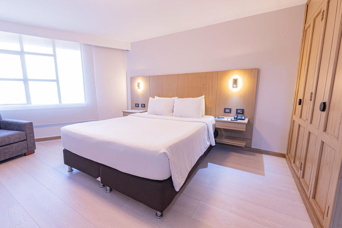 A hotel room with a large bed, bedside tables, a desk, a chair, and a wardrobe.