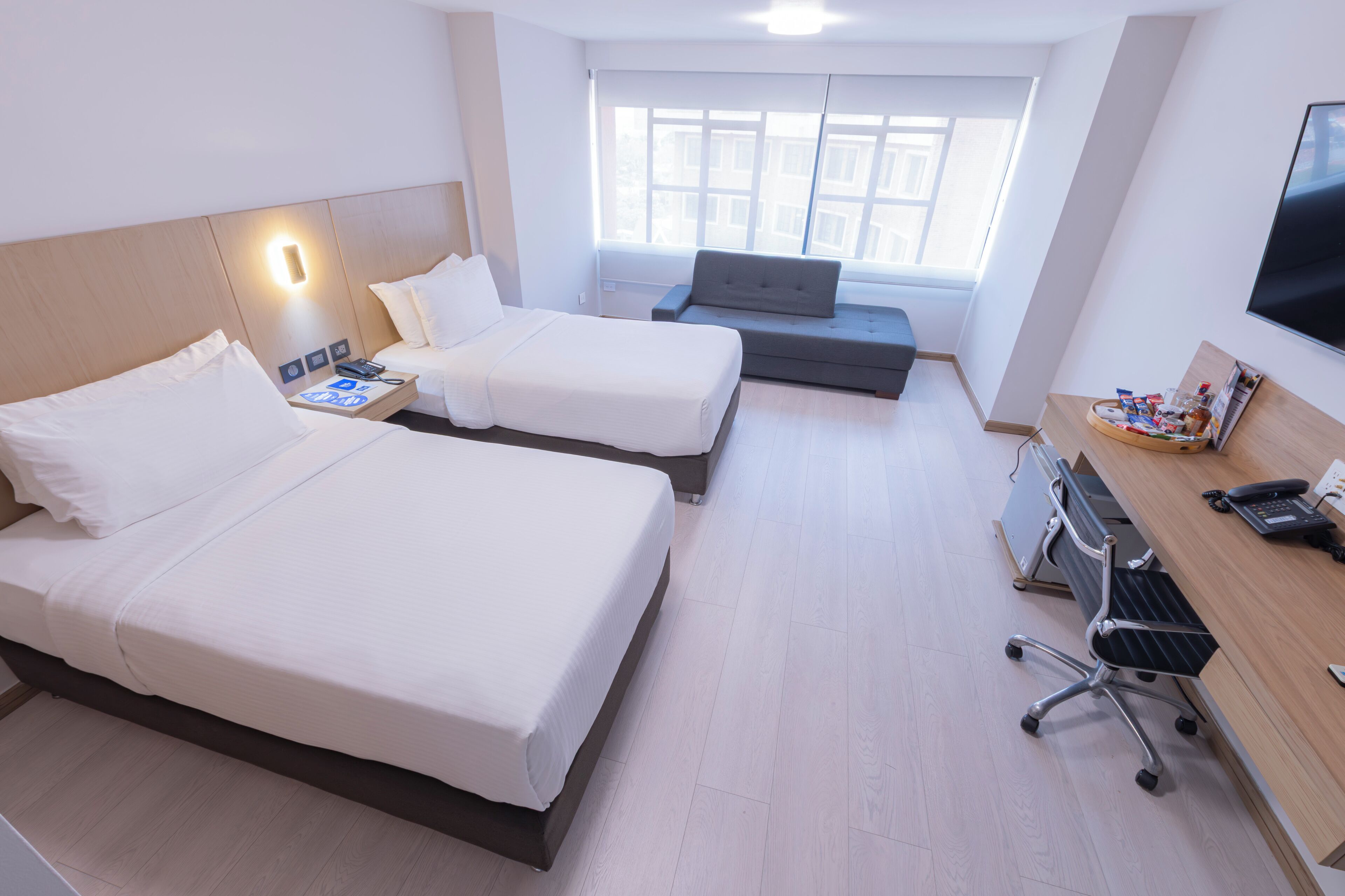Habitación de hotel con dos camas, un escritorio con computadora, televisor y ventana con vista a edificios.