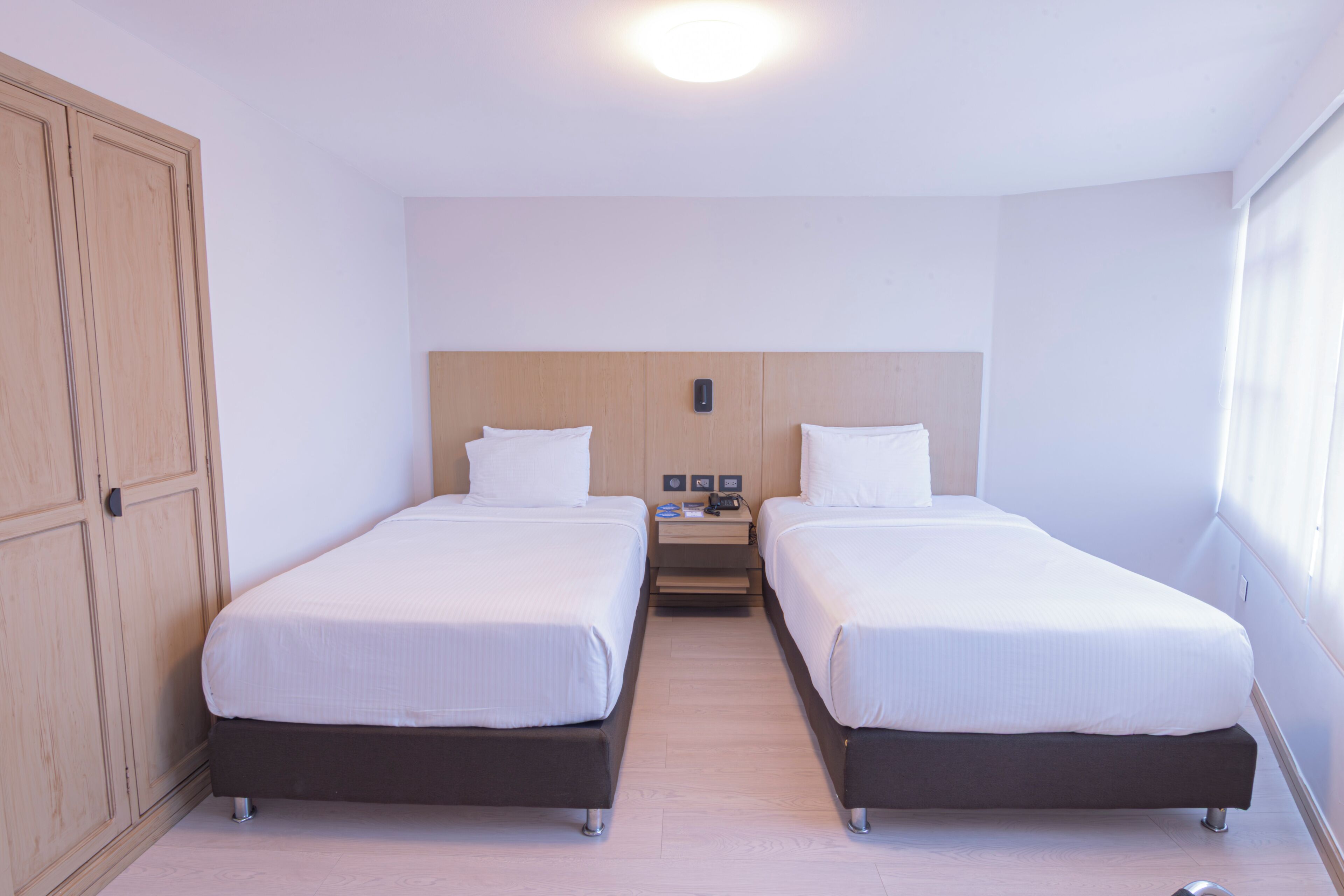 A hotel room with two single beds, a wooden wardrobe, a nightstand with a lamp, and a window with curtains.