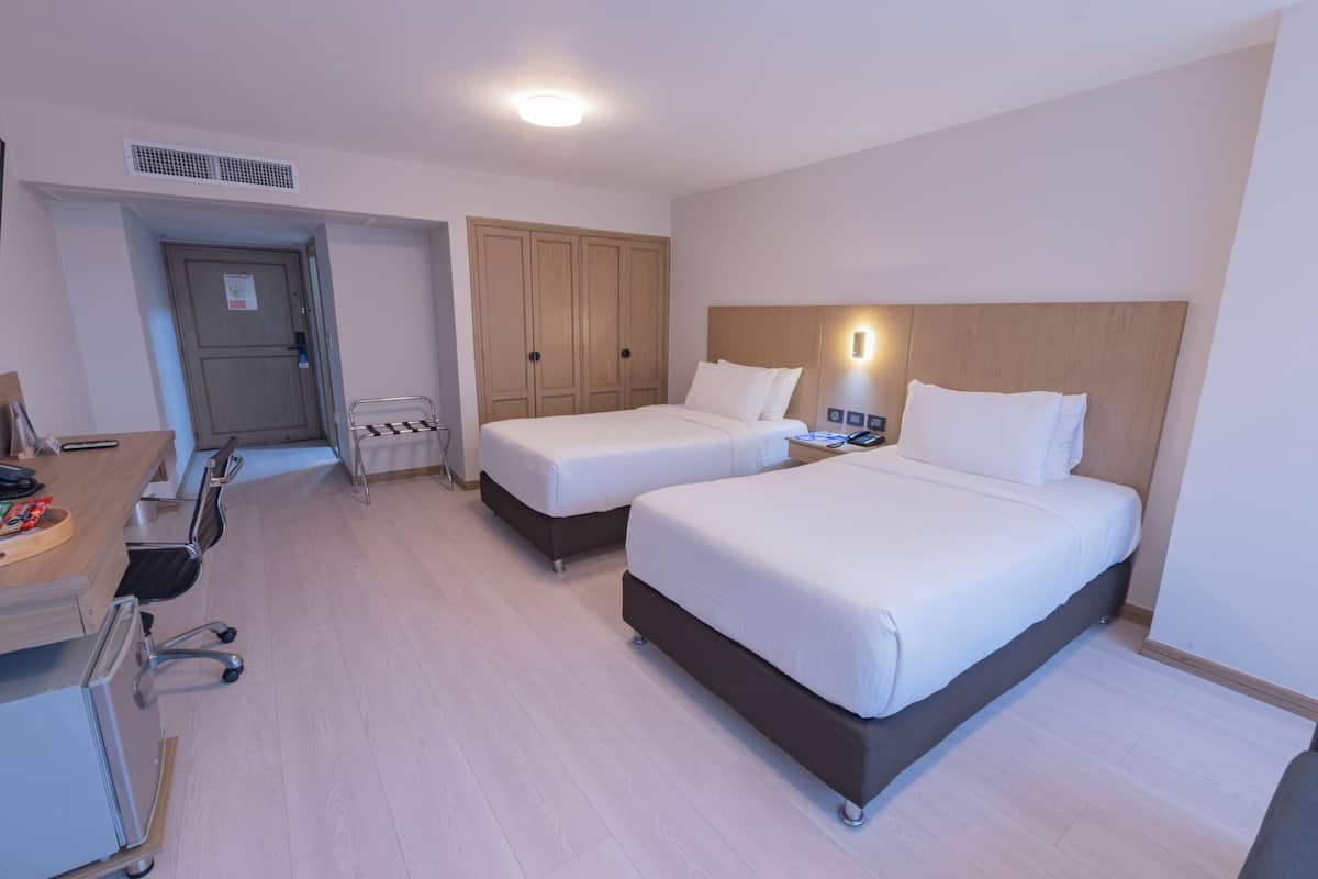 A hotel room with two beds, a desk with a chair, a wardrobe, and a bedside table with a lamp.