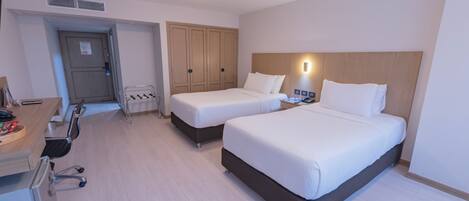 Quarto de hotel com duas camas, uma escrivaninha com cadeira, um guarda-roupa e uma mesa de cabeceira com abajur.