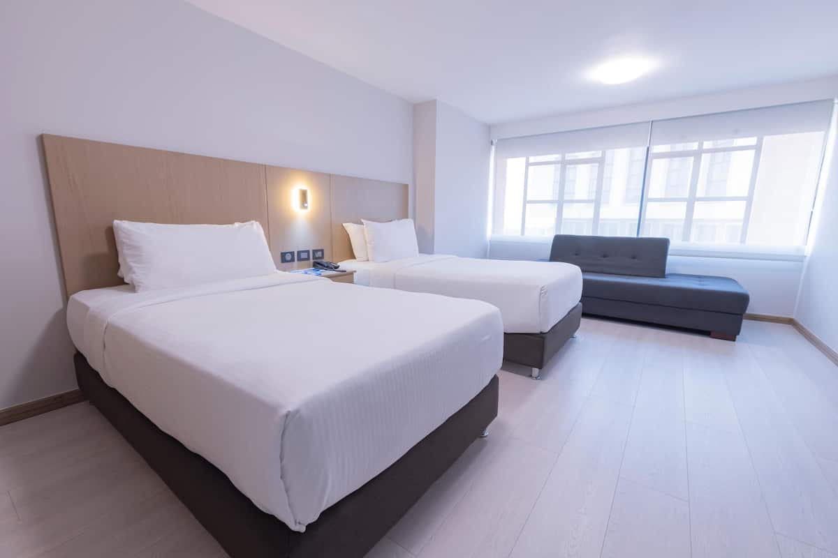 A hotel room with two beds, a sofa, a bedside table with a lamp, and a large window.