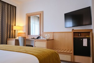 Habitación de hotel con cama, escritorio, silla, lámpara, espejo, televisor y refrigerador.