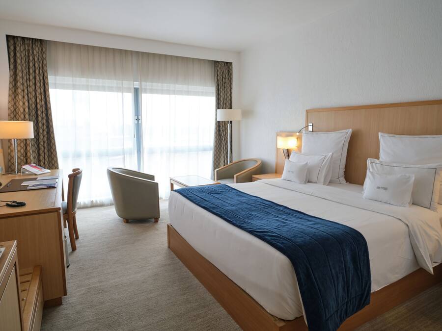 A hotel room with a large bed, a desk with a chair, a lamp, and a view of the outdoors through the window.