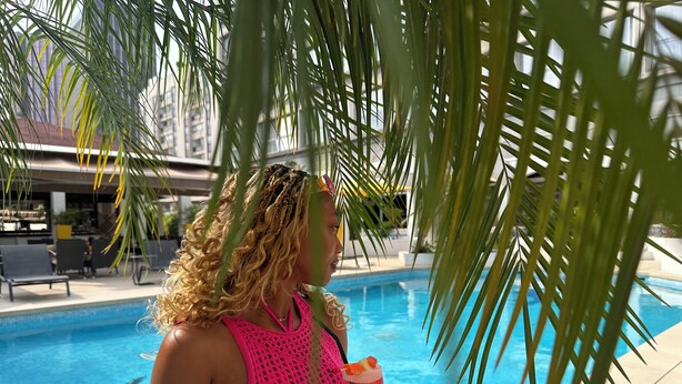 Une personne vêtue d’un maillot de bain rose, tenant un verre près d’une piscine, avec des palmiers et des chaises longues en arrière-plan.