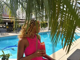 Una persona con traje de baño rosa sosteniendo una bebida junto a una piscina, con palmeras y sillones de descanso al fondo.