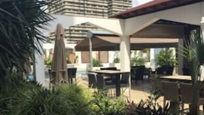 Un espace patio sur le toit, doté de sièges extérieurs et de parasols, entouré de verdure et avec un grand bâtiment en arrière-plan.
