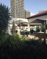 Un patio en la azotea con mobiliario exterior y sombrillas, rodeado de vegetación y un edificio alto al fondo.