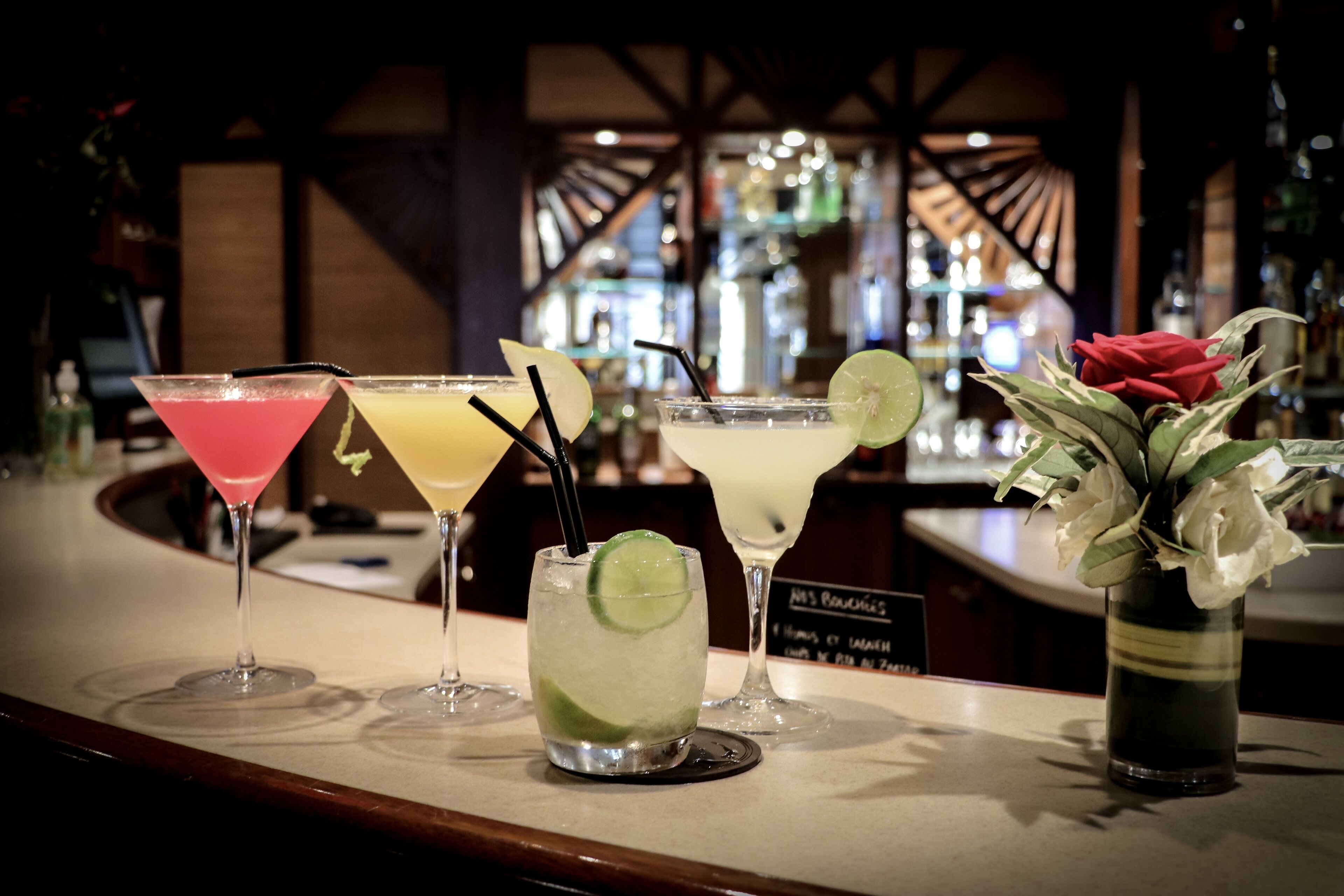 A bar counter with various cocktails, a vase with flowers, and a bar sign.
