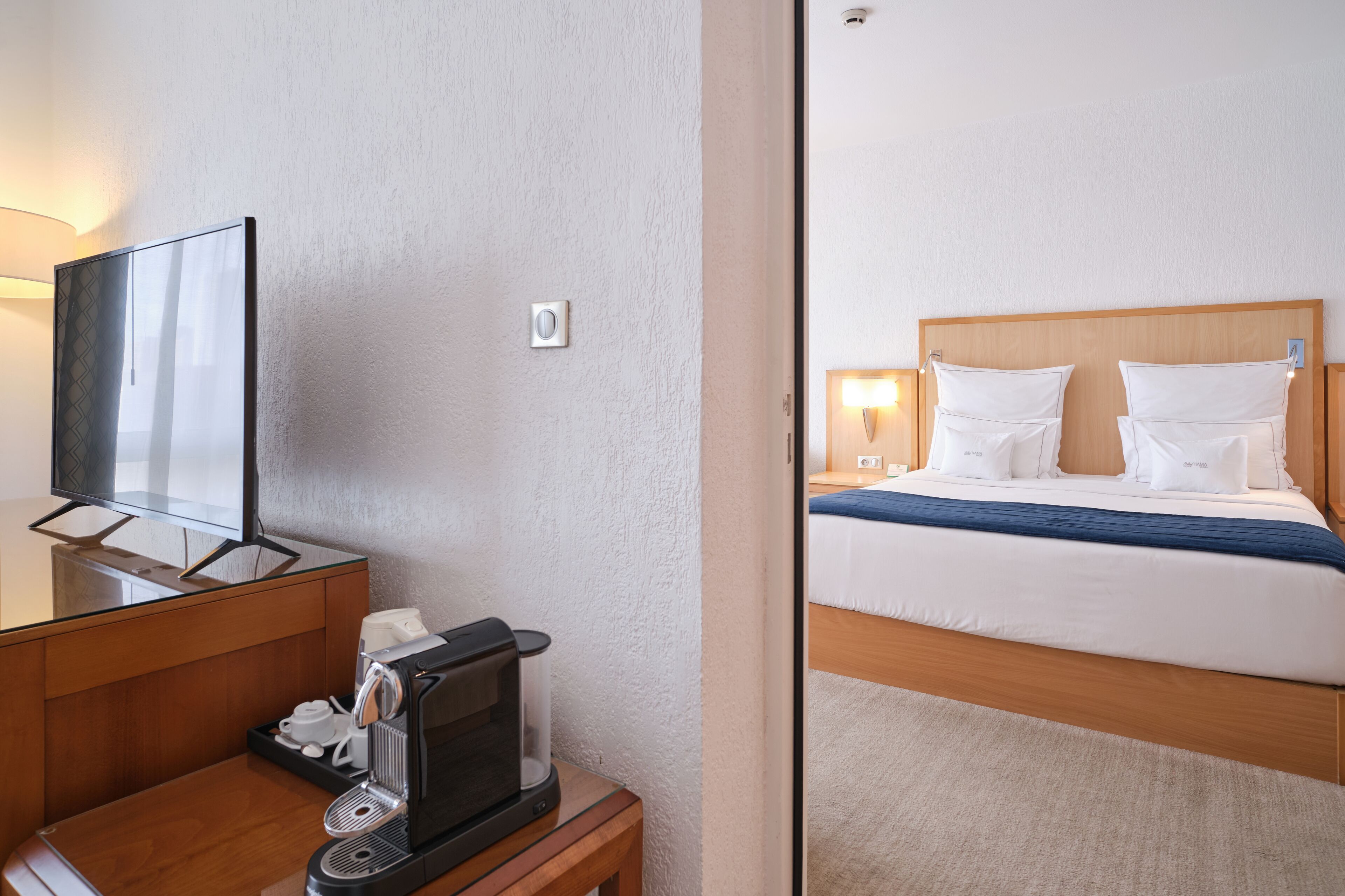 Una habitación de hotel con un escritorio de madera, una cafetera, un televisor y una cama con ropa de cama blanca y una manta azul.