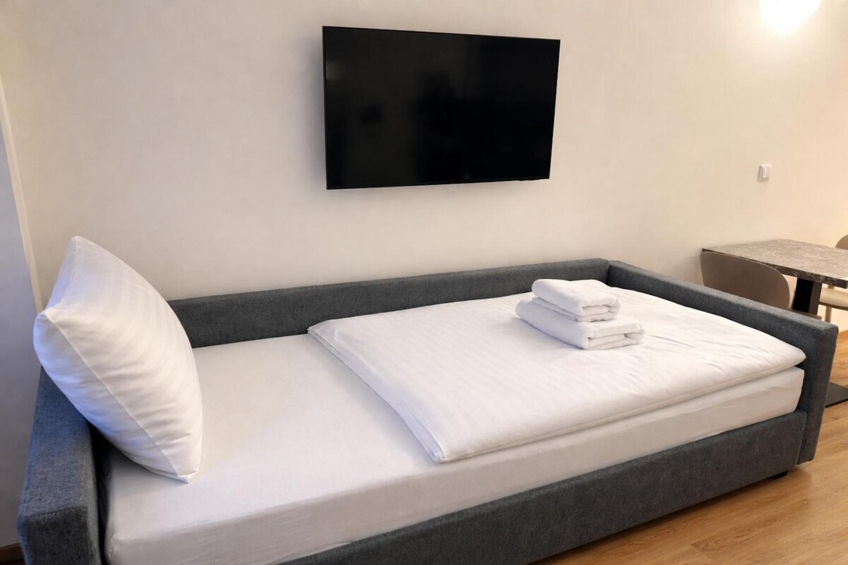 A single-bed room with a grey sofa bed, a white pillow, and a folded white towel on the bed.