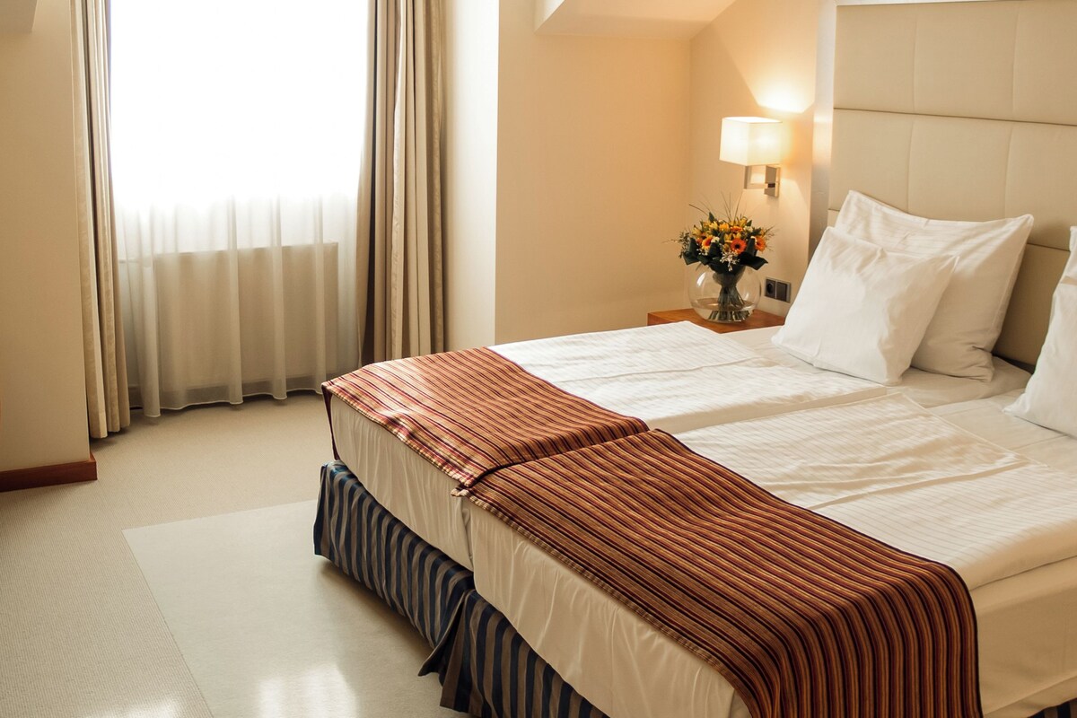 A hotel room with a large bed, a nightstand with a lamp, a window with curtains, and a ceiling light.