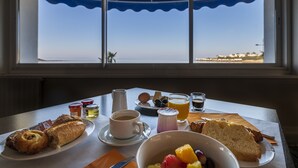 A breakfast table with croissants, a bowl of fruit, coffee, and juice, viewed through a window overlooking a beach. - CERISE Royan - Le Grand Hôtel de la Plage (Royan)