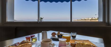 Una mesa de desayuno con croissants, un bol de fruta, café y zumo, vista a través de una ventana con vistas a una playa.