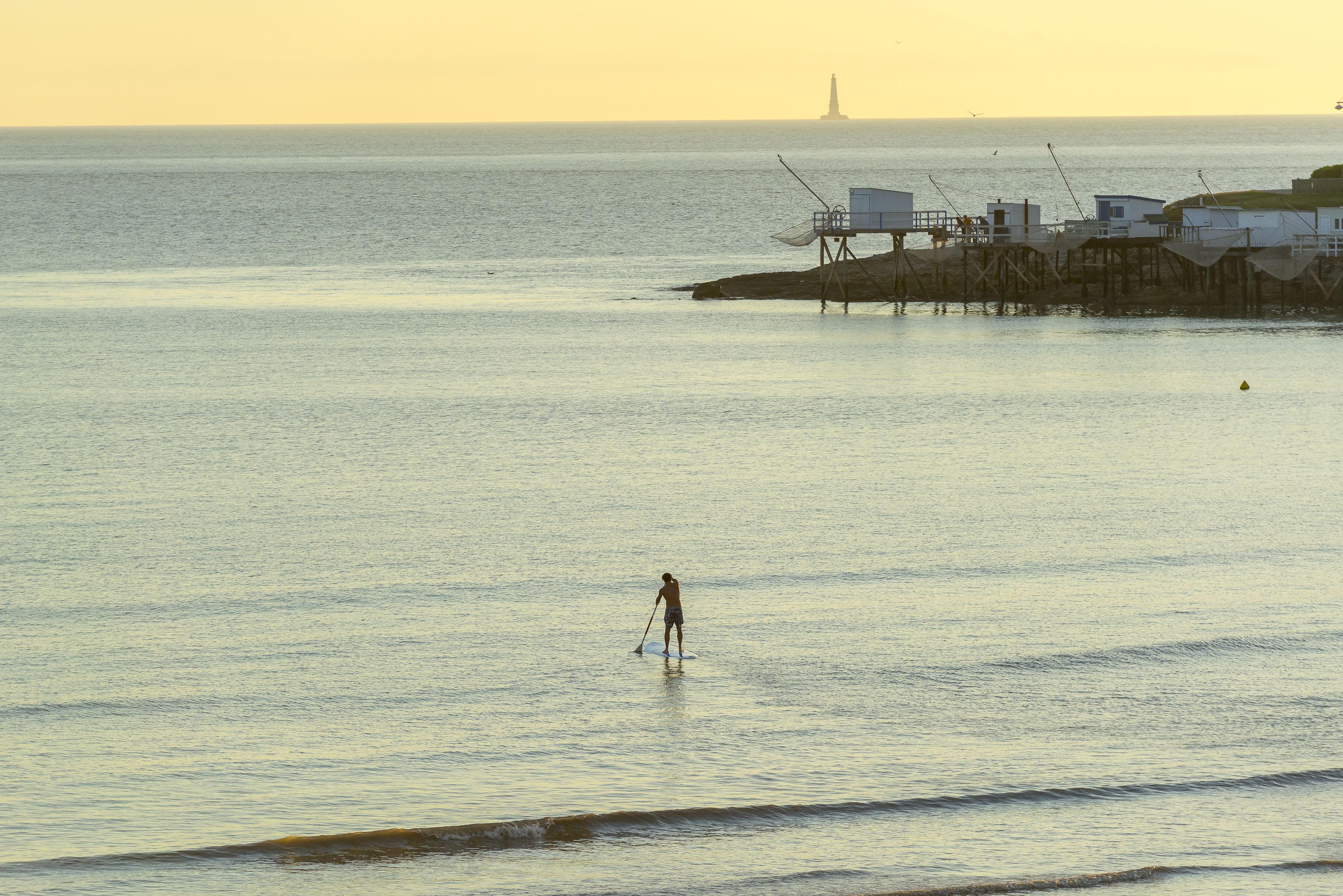 A person paddleboarding on calm water with a lighthouse in the distance.