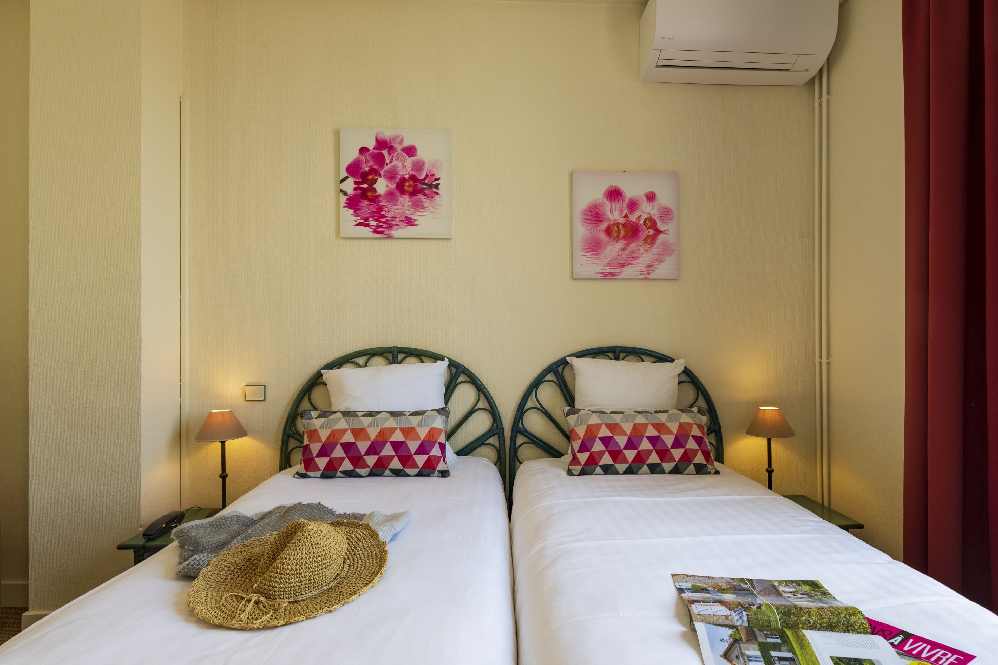 Une chambre d’hôtel avec deux lits, chacun agrémenté d’un oreiller à motifs et d’une lampe. On y trouve deux climatiseurs fixés au mur et deux œuvres d’art florales encadrées.