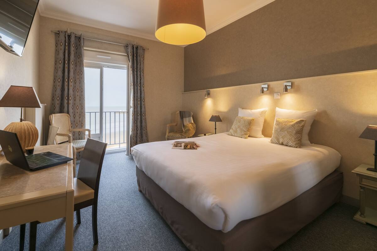 A hotel room with a large bed, a desk with a laptop, a chair, and a balcony with curtains.