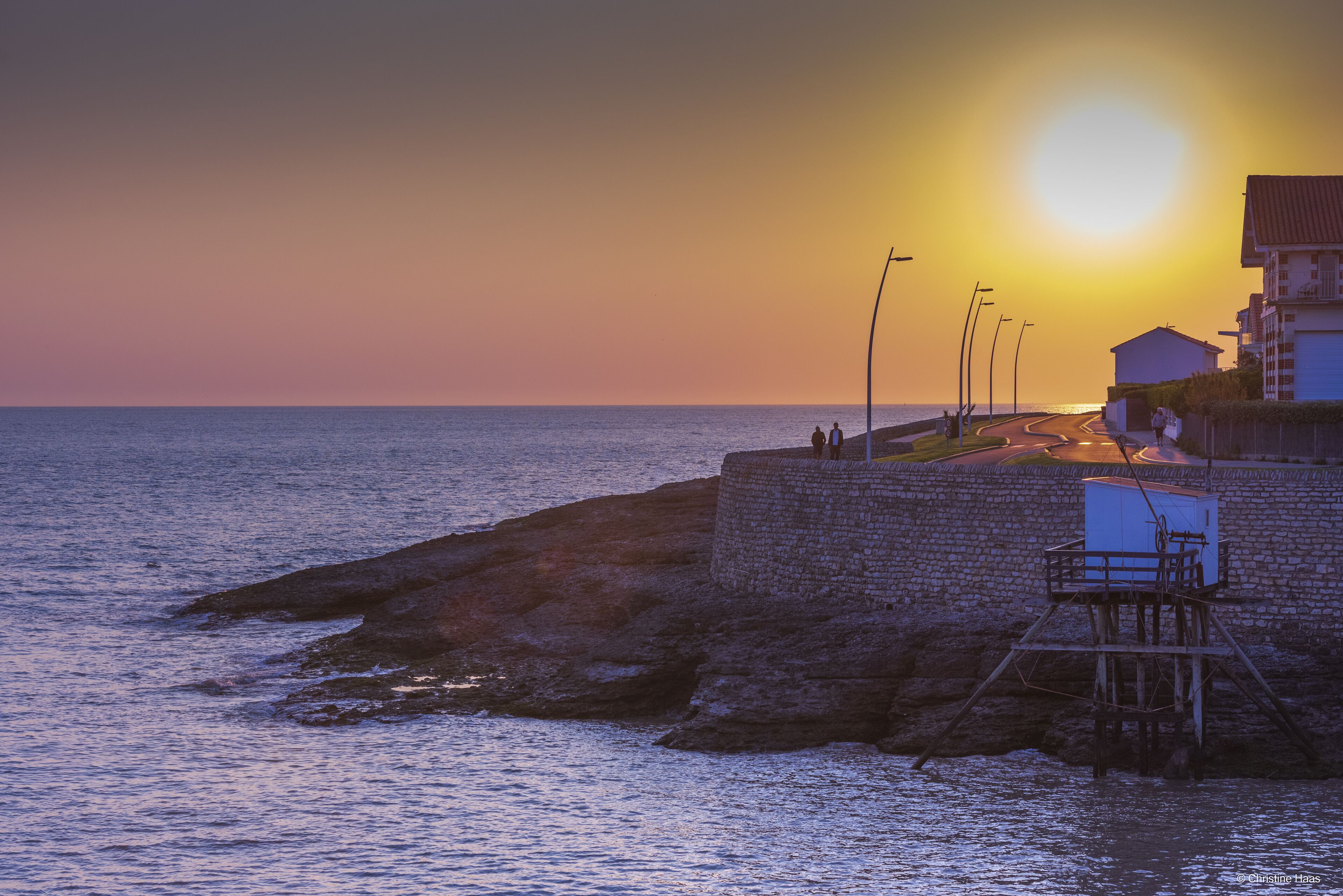 Escena costera con un atardecer, un muro de piedra y un edificio con una escalera que conduce a una plataforma.