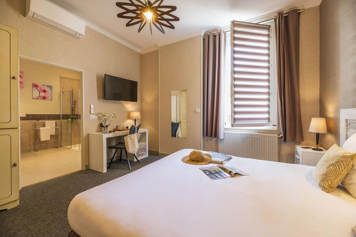 A hotel room with a bed, desk, shower, TV, and a window with curtains.