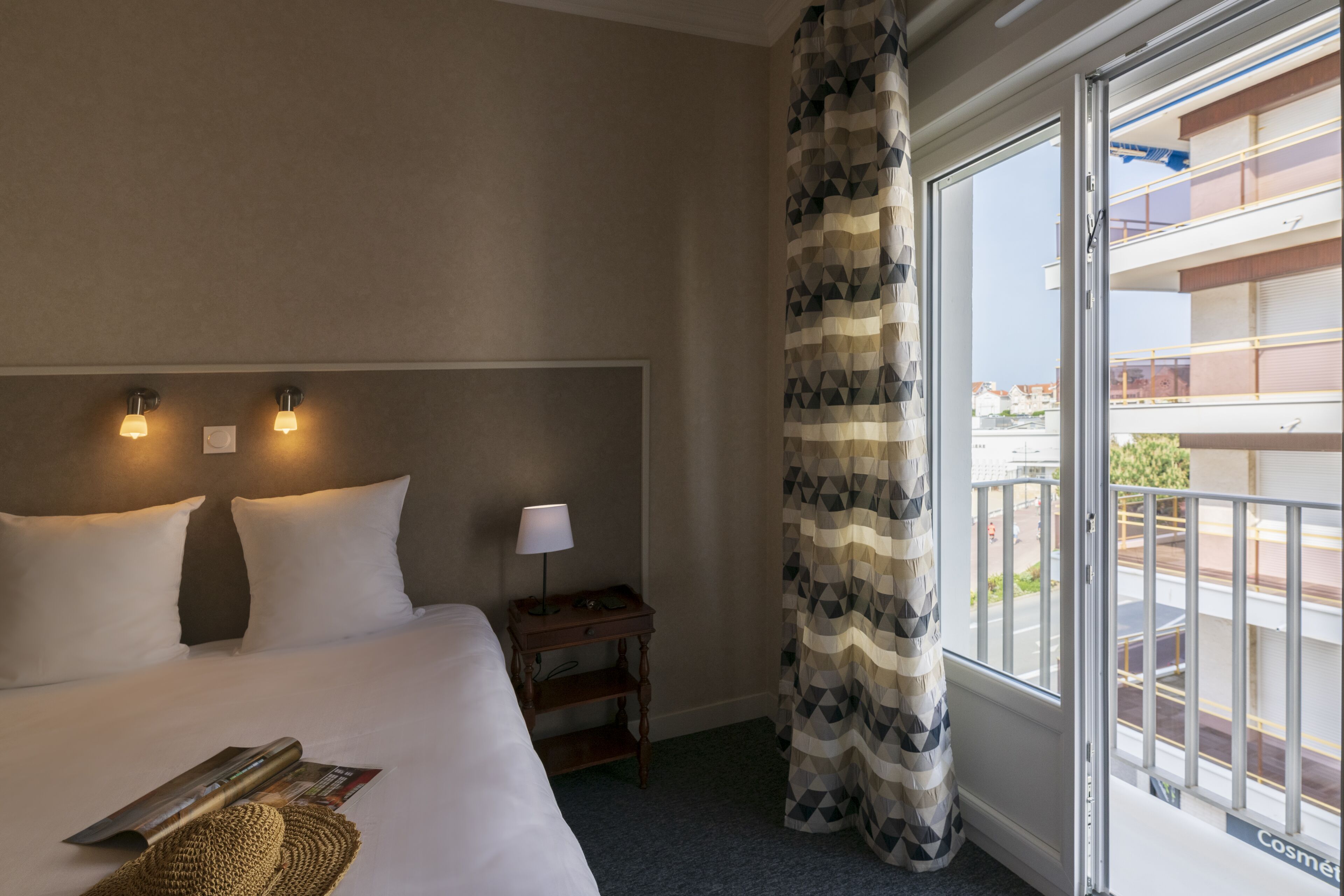 Une chambre d’hôtel avec un grand lit, deux lampes de chevet, une table de chevet avec une lampe, une fenêtre donnant sur l’extérieur et un rideau à motifs.
