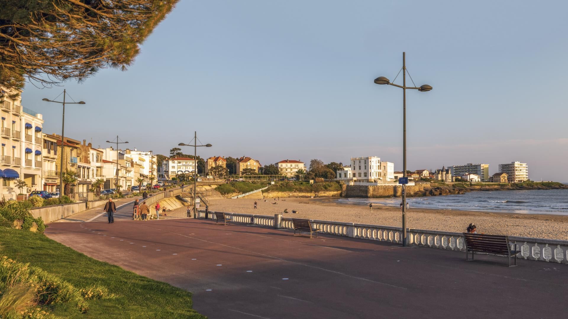 A coastal promenade with a paved walkway, benches, street lamps, and buildings in the background.