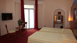 A hotel room with a bed, a desk, a chair, a TV, a plant, a fireplace, and a window with red curtains. - Inter Hotel Bristol (Aix-les-Bains)