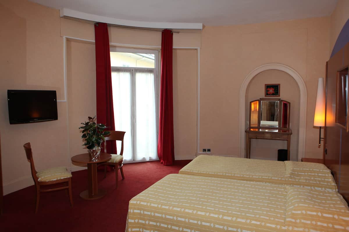 A hotel room with a bed, a desk, a chair, a TV, a plant, a fireplace, and a window with red curtains.