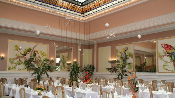 A formal dining room with multiple tables set for guests, featuring a high ceiling with decorative lighting and large floral paintings on the walls. - Inter Hotel Bristol (Aix-les-Bains)