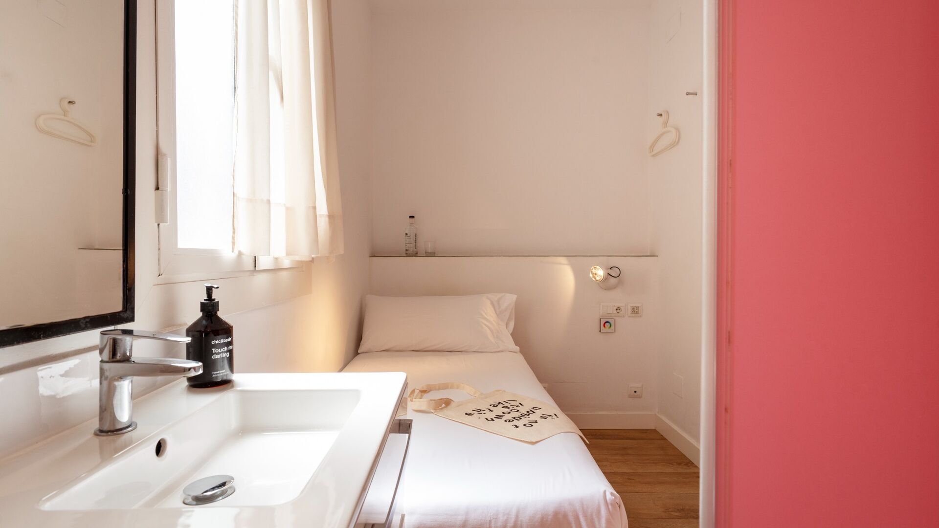 Une petite pièce propre, avec un lavabo blanc, un miroir, une bouteille de shampoing et un lit avec un oreiller blanc.