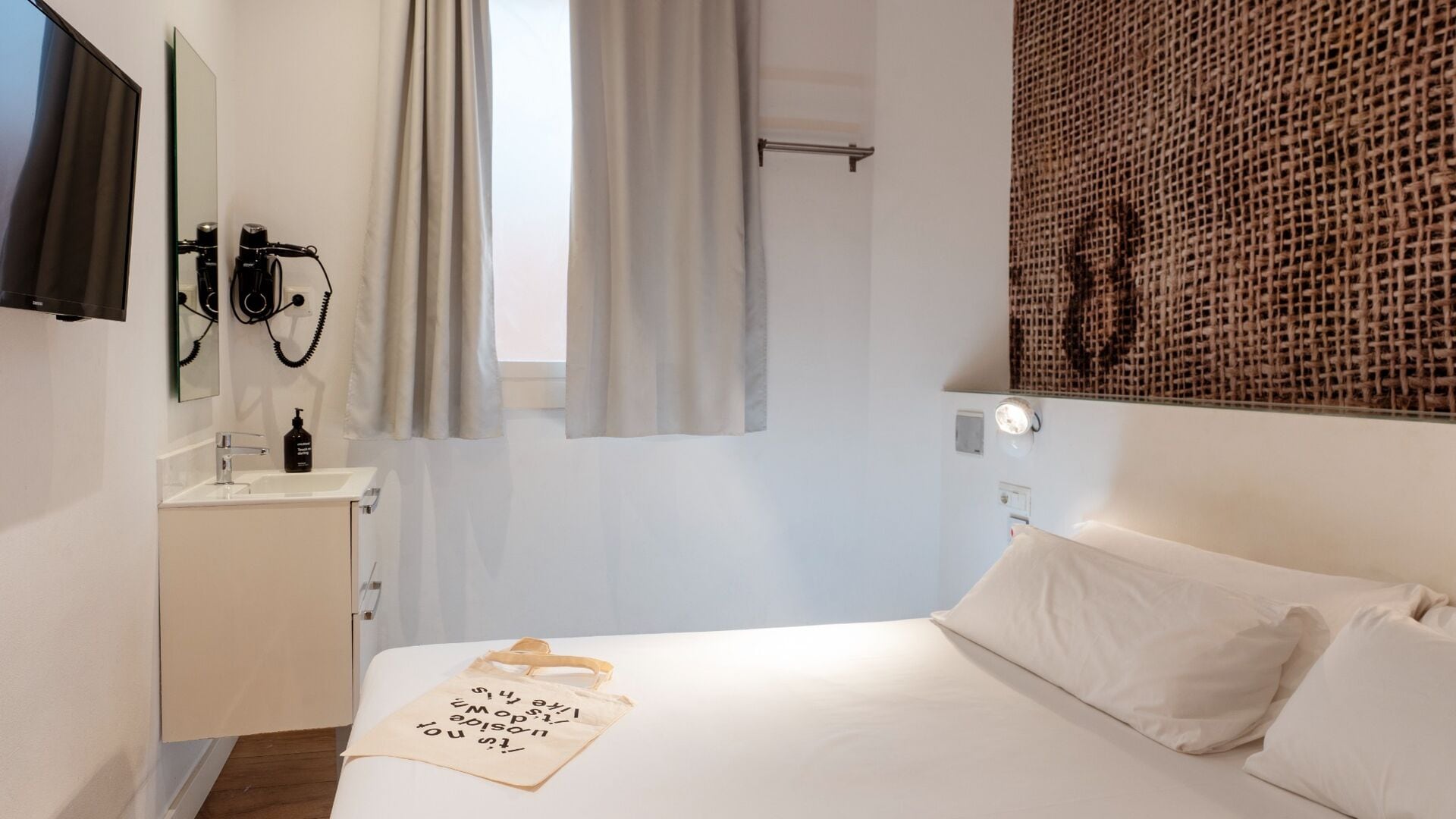 A bedroom with a bed, a bedside table with a bottle, a mirror, and a wall-mounted light fixture.