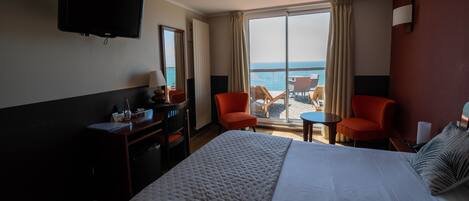 A hotel room with a large bed, a desk with a lamp, a TV, and a view of the ocean through the window.