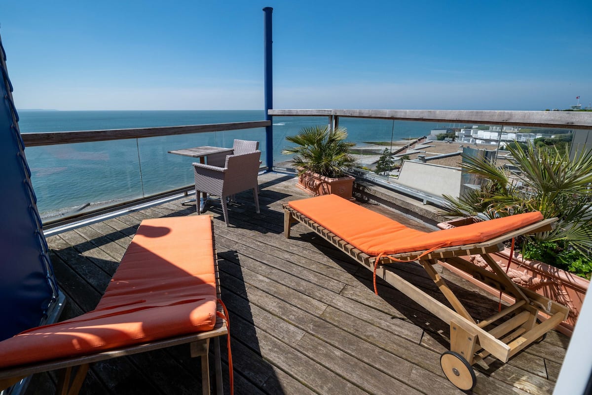 Terraza de madera con dos sillones de descanso y una mesa, con vista a una playa y el océano.