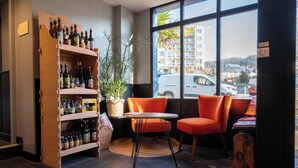 A cozy seating area with a wooden shelf filled with bottles, a potted plant, and a view of the city through large windows.